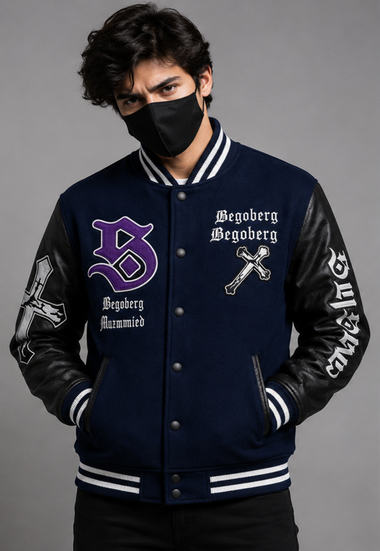 A man wearing a navy blue varsity-style jacket with black leather sleeves, white striped ribbing, and gothic embroidery reading 'Begoberg' and 'Hannibal' on a gray studio background.