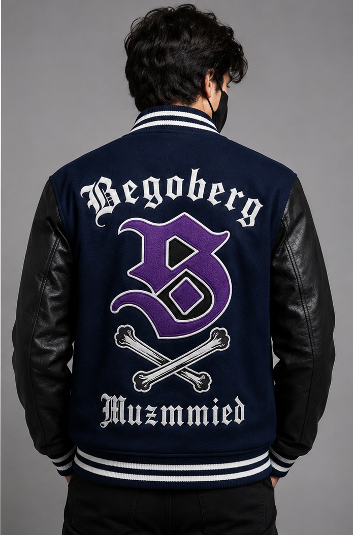 A man viewed from behind wearing a navy blue varsity-style jacket with black leather sleeves. The back features white gothic lettering reading 'Begoberg' and 'Muzmmied' with a large purple letter 'B' and crossed bones in the center.