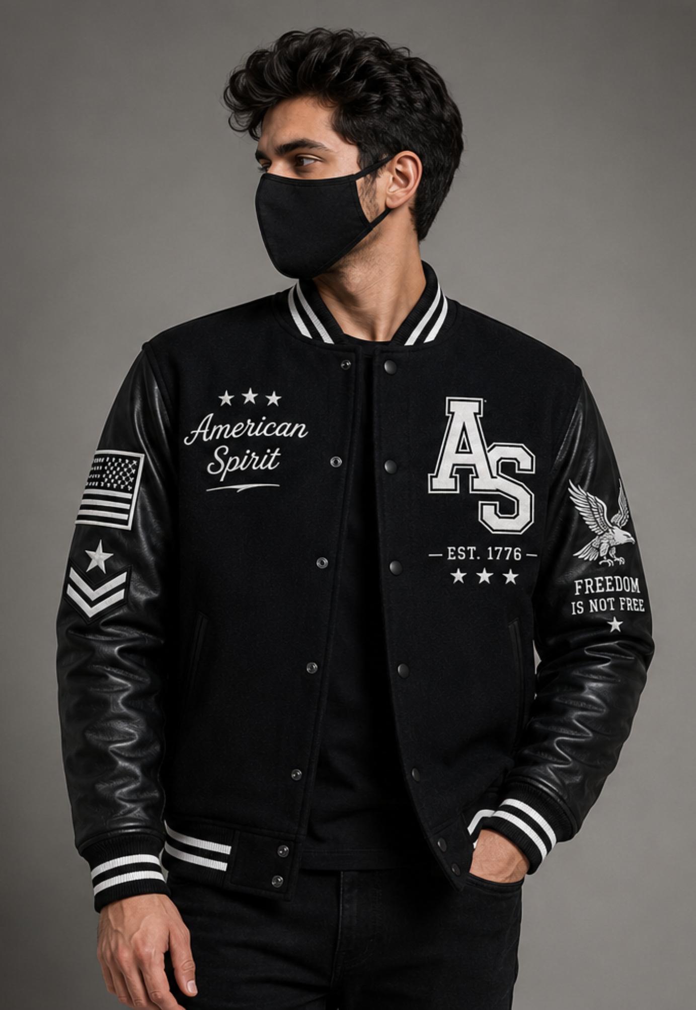 A man wearing a black varsity-style bomber jacket with white embroidered logos, including 'American Spirit' on the left chest and 'AS EST. 1776' on the right chest, paired with a black face mask against a dark gray studio background.