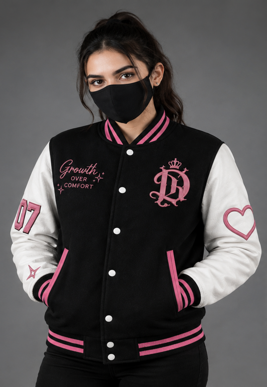 A woman wearing a black and white varsity jacket with pink accents, featuring an embroidered pink monogram logo and the words 'Growth OVER COMFORT' on the chest, posed against a solid gray studio background.