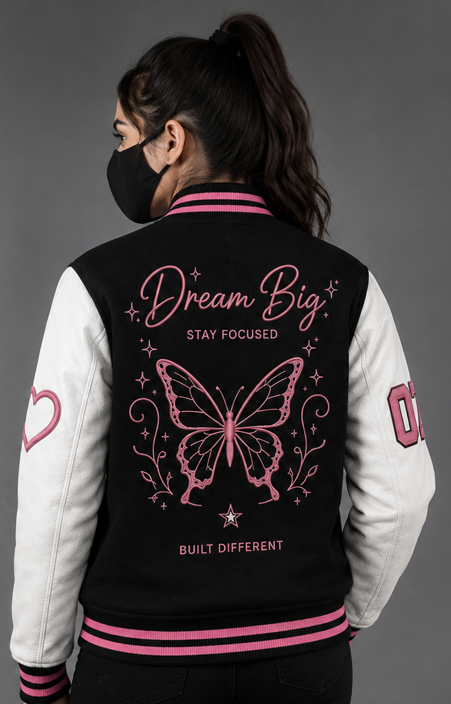 A woman shown from the back wearing a varsity-style jacket with a black body, white sleeves, and pink trim. The back features a large pink butterfly graphic with the words 'Dream Big', 'STAY FOCUSED', and 'BUILT DIFFERENT'.