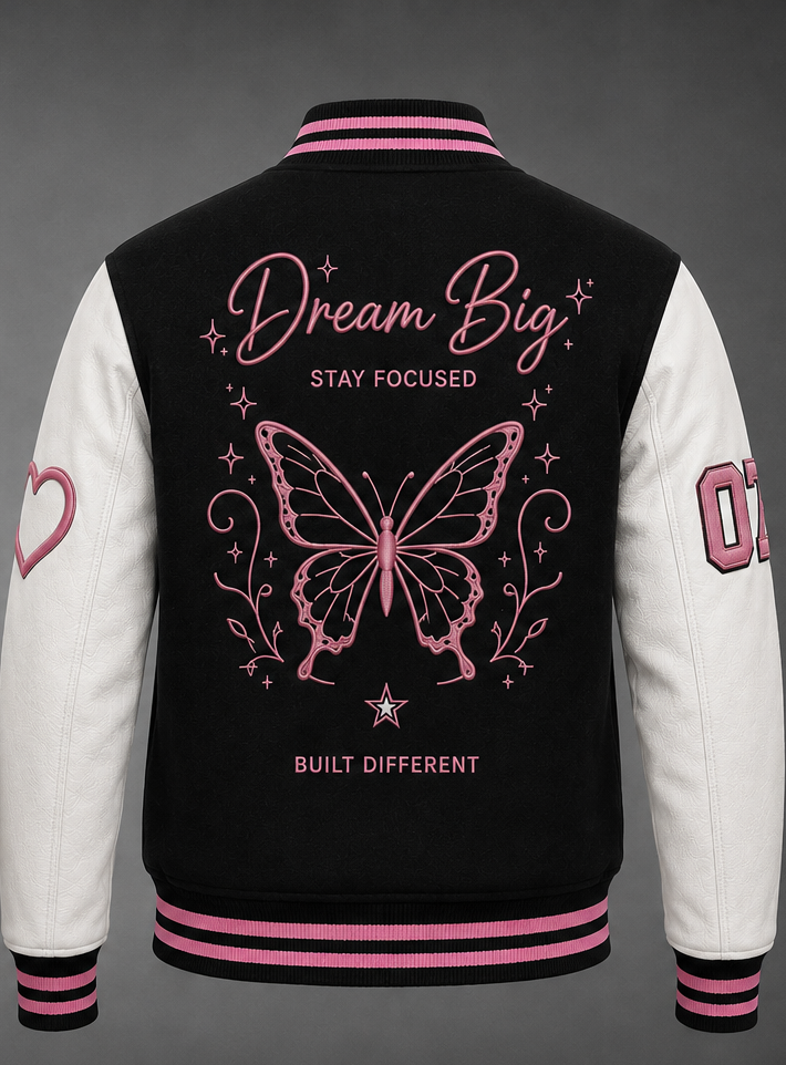 Black and white varsity jacket shown from the back, featuring pink and white graphic designs including a butterfly, 'Dream Big' text, and pink striped ribbed collar, cuffs, and hem.