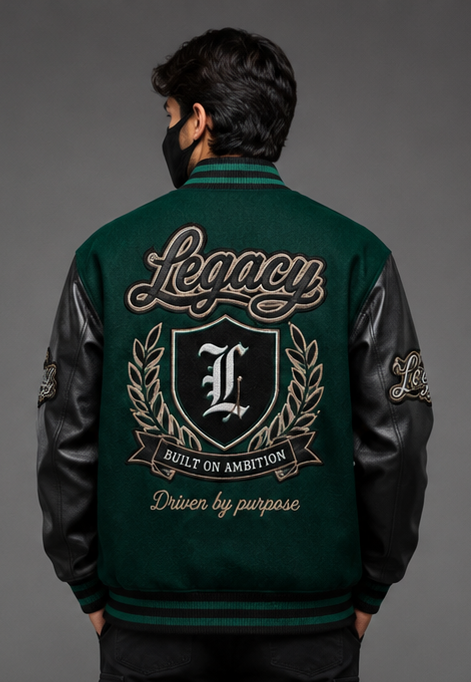 A man viewed from behind wearing a dark green and black varsity-style jacket with a large embroidered 'Legacy' crest and 'Built on Ambition' text on the back, against a dark gray studio background.