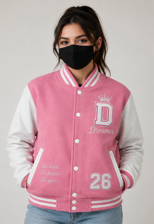 A woman wearing a pink and white varsity jacket with a black face mask, shown from the waist up against a light gray studio background.