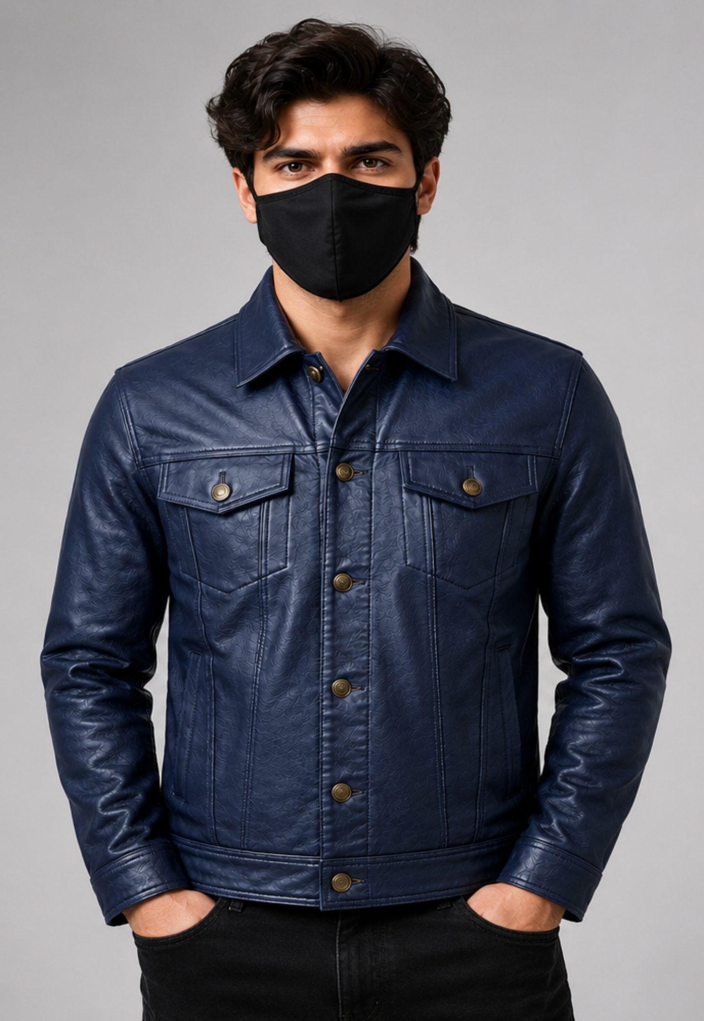 A man wearing a dark navy blue leather trucker jacket with brown buttons, paired with a black face mask and black pants, standing against a solid gray studio background.