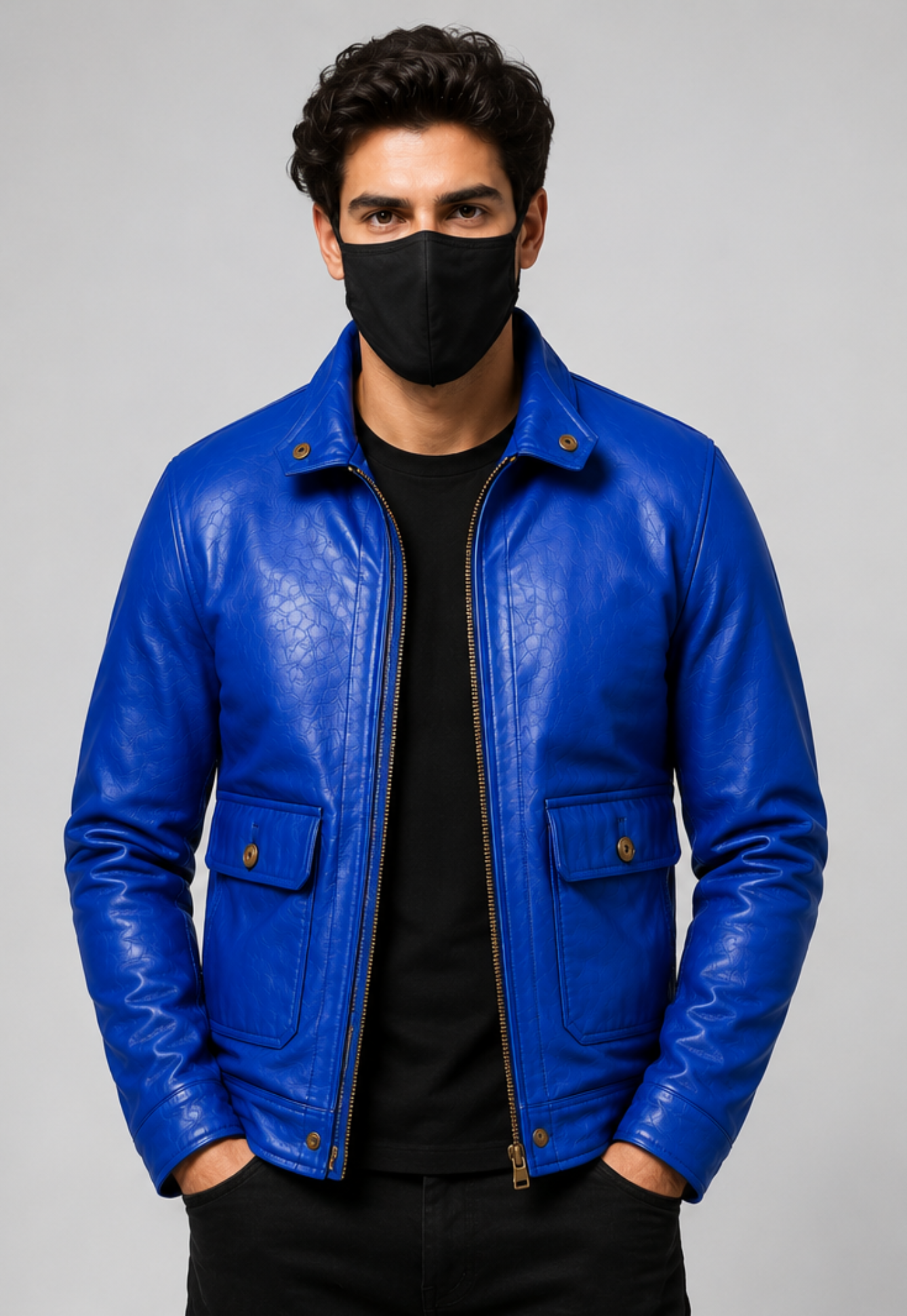 A man wearing a vibrant blue leather jacket over a black t-shirt and black pants, with a black face mask, photographed against a neutral gray studio background.