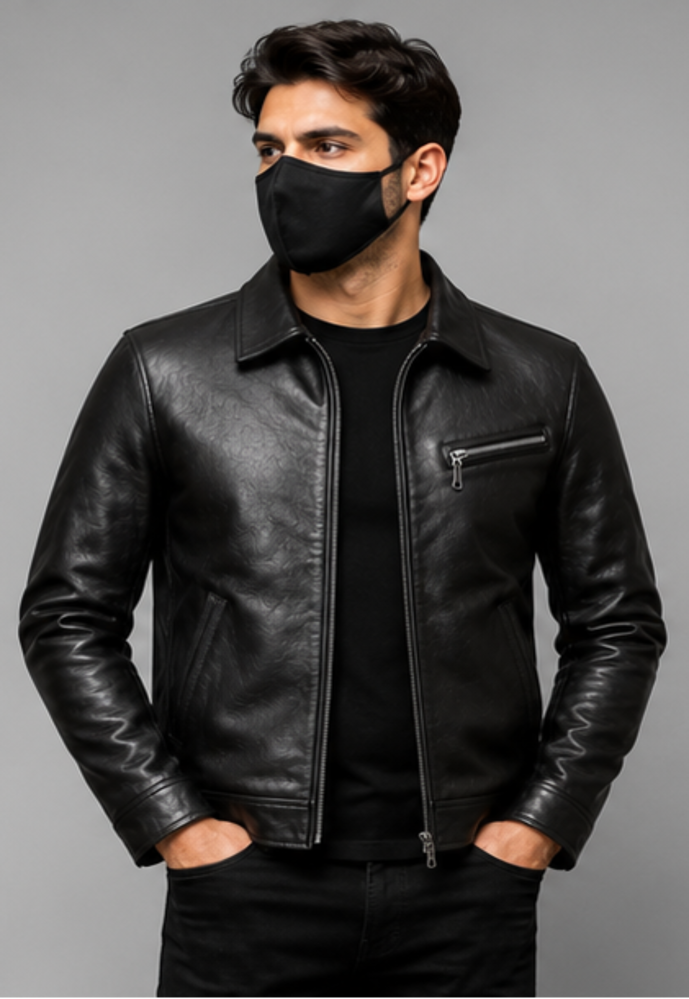 A man wearing a black leather jacket over a black t-shirt and black pants, with a black face mask, standing against a solid gray studio background.