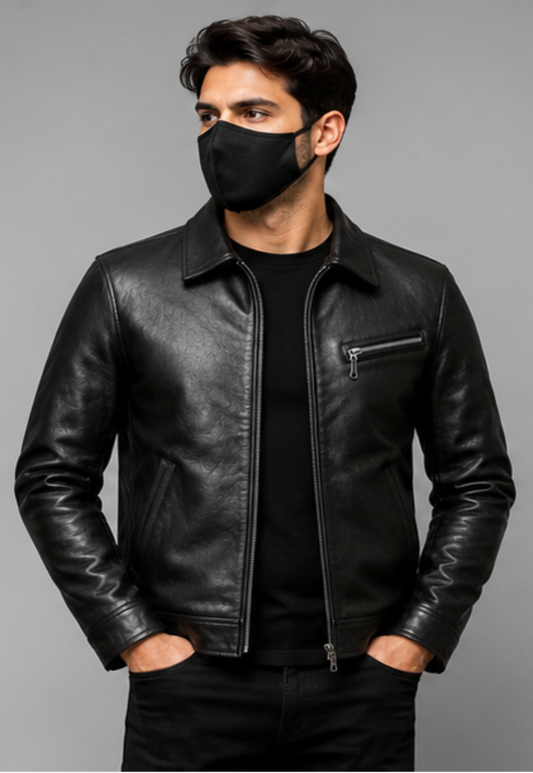 A man wearing a black leather jacket over a black t-shirt and black pants, with a black face mask, standing against a solid gray studio background.