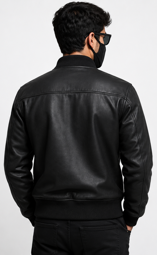 A man viewed from the back wearing a black leather bomber jacket, black pants, a black face mask, and black sunglasses against a light gray studio background.