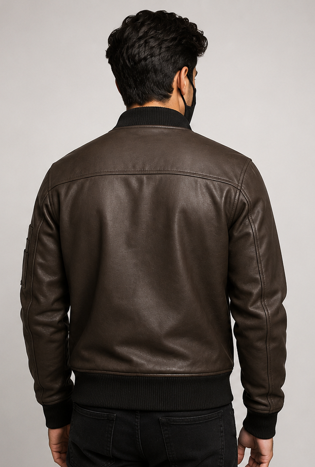 A man viewed from the back wearing a dark brown leather bomber jacket with black ribbed trim at the collar, cuffs, and hem, paired with black jeans against a light gray studio background.
