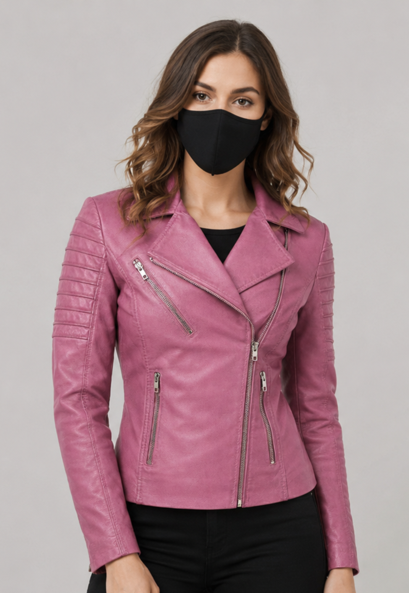 A woman wearing a pink leather moto jacket with silver zippers, styled with a black face mask and black pants, against a solid gray studio background.