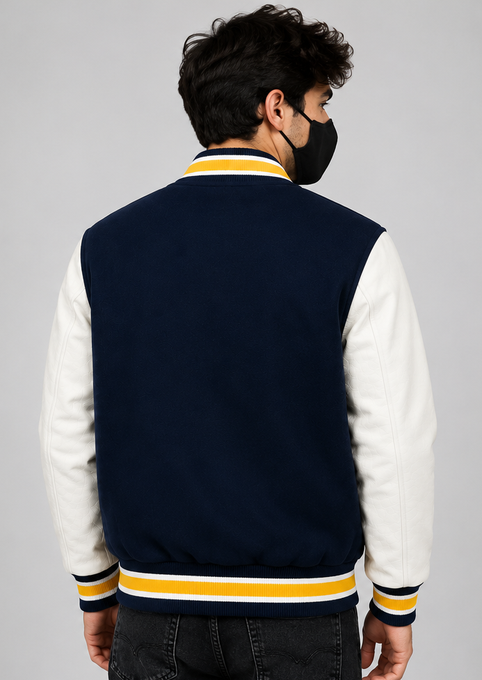 A man viewed from the back wearing a navy blue and white varsity jacket with yellow stripes on the collar, cuffs, and hem, paired with a black face mask and dark jeans, against a solid gray studio background.