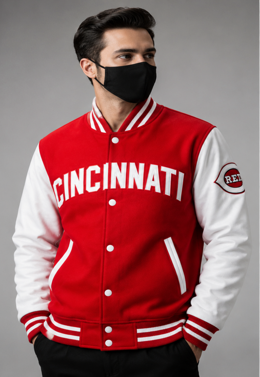 A man wearing a red and white Cincinnati varsity jacket with 'CINCINNATI' in white letters across the chest, paired with a black face mask, against a solid gray studio background.