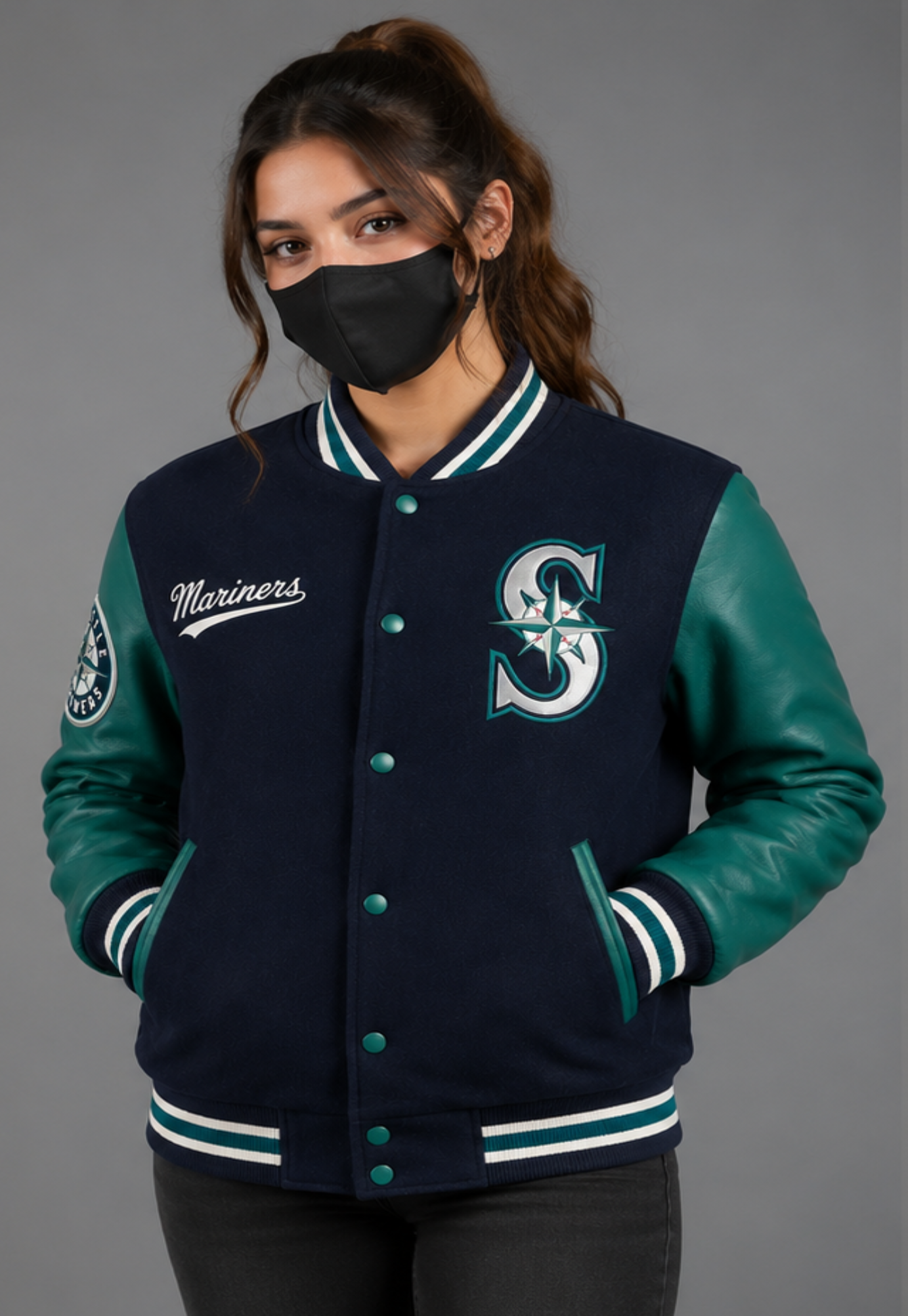 A woman wearing a Seattle Mariners varsity jacket with navy blue wool body, teal leather sleeves, and a black face mask, photographed against a solid gray studio background.