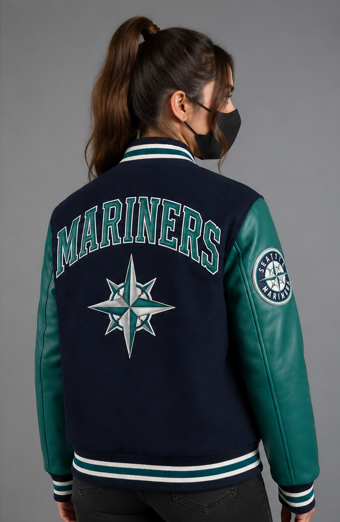 A woman viewed from the back wearing a Seattle Mariners varsity jacket with navy blue body, teal leather sleeves, and the team name and logo on the back, against a plain gray studio background.