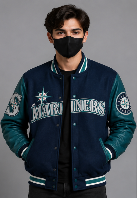 A man wearing a Seattle Mariners varsity jacket with navy blue and teal leather-like panels, white lettering, and a black face mask, photographed against a plain gray studio background.