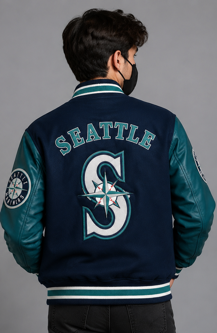 A man viewed from behind wearing a Seattle Mariners varsity jacket. The jacket features a navy blue body with teal sleeves and collar, and the Seattle Mariners 'S' logo and 'SEATTLE' text embroidered on the back.