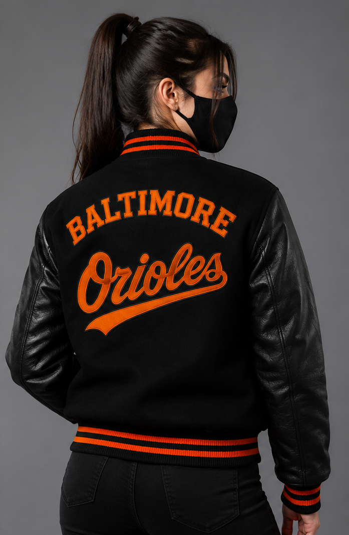 A woman shown from the back wearing a black varsity-style jacket with orange lettering that reads 'BALTIMORE Orioles' on the back, black leather-look sleeves, and orange striped ribbing, against a dark gray studio background.