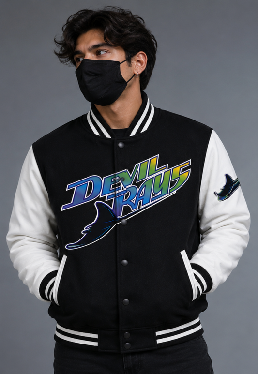 A man wearing a black-and-white varsity jacket with a colorful 'Devil Rays' graphic embroidery on the chest, paired with a black face mask, set against a solid gray studio background.