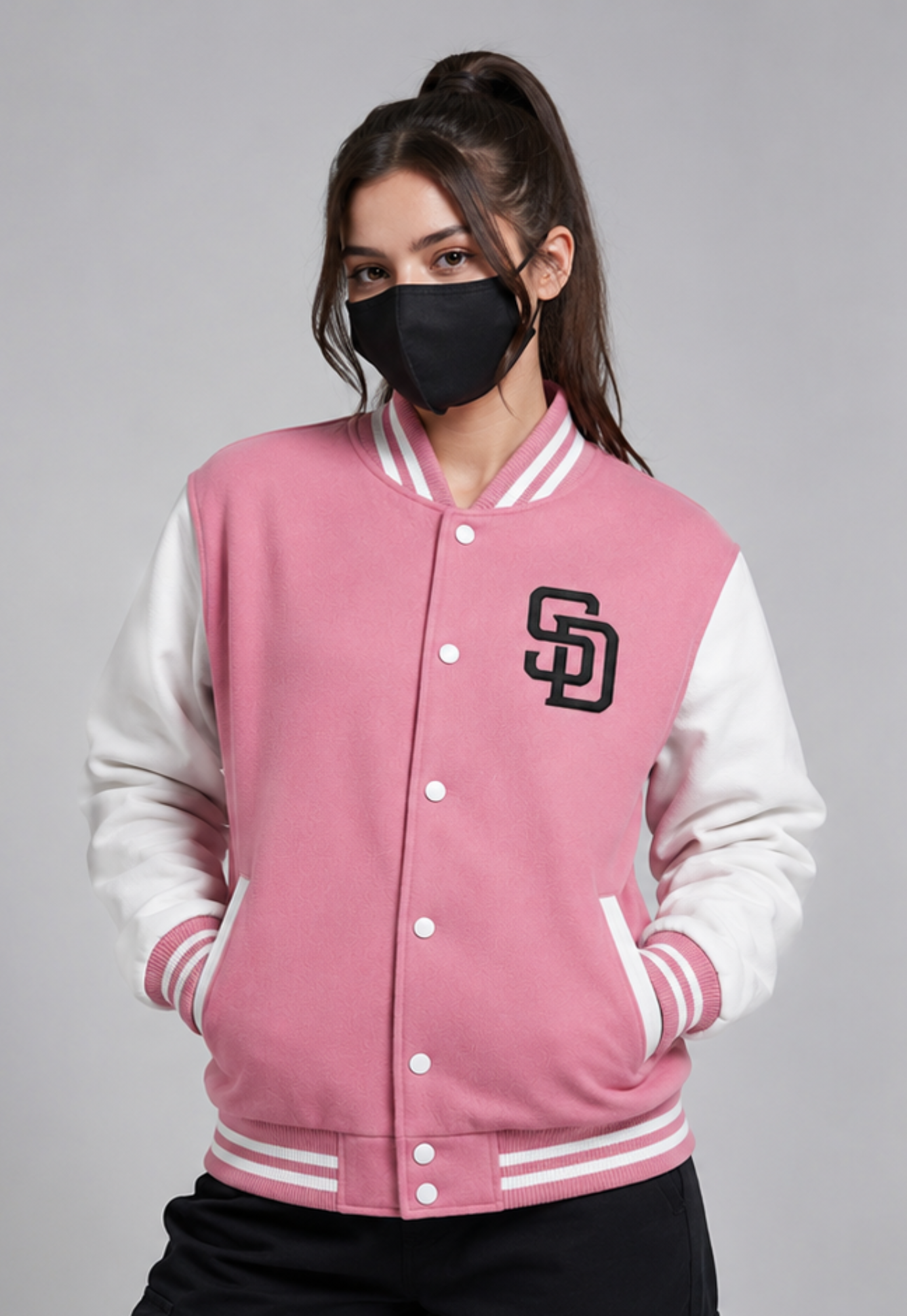 A woman wearing a pink and white varsity jacket with a black embroidered 'SD' logo on the chest, posing against a solid gray studio background.