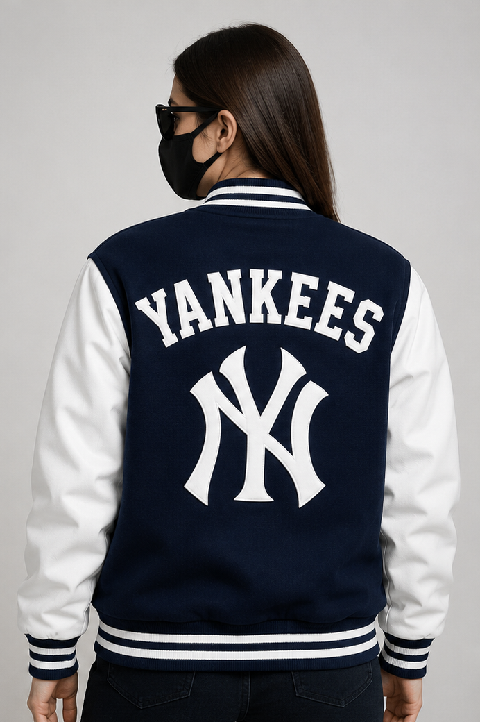 A woman shown from the back wearing a navy and white varsity-style jacket with large white 'YANKEES' lettering and the New York Yankees 'NY' logo on the back, photographed against a plain light gray studio background.