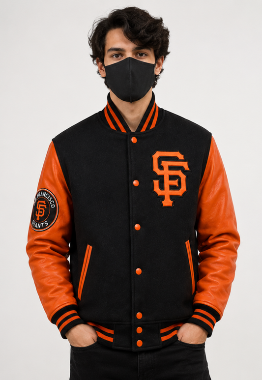A man wearing a black and orange San Francisco Giants varsity jacket, a black face mask, and black pants, standing against a light gray studio background.