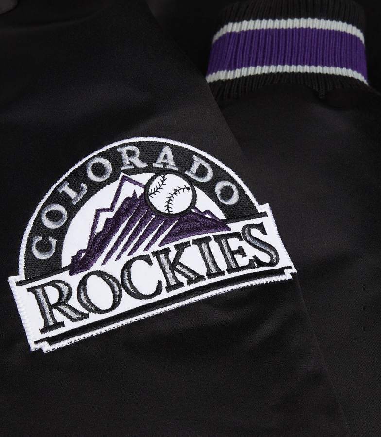 Close-up of a black Colorado Rockies baseball jacket featuring a large embroidered team logo patch on the chest and a purple and white striped ribbed collar.