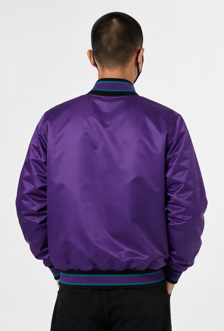A man viewed from behind wearing a purple satin bomber jacket with dark green and navy striped ribbed collar, cuffs, and hem, paired with black pants against a light gray background.