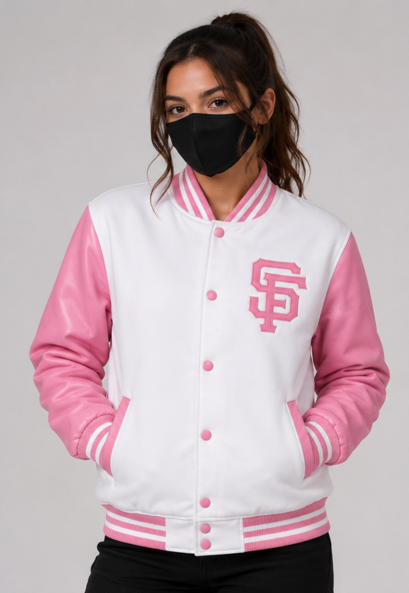 A woman wearing a pink and white varsity jacket with a pink 'SF' logo on the chest, paired with a black face mask and black pants, against a light gray studio background.