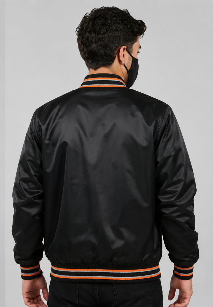 A man viewed from the back wearing a black satin bomber jacket with orange and navy striped ribbed collar, cuffs, and hem, paired with a black face mask against a light gray studio background.