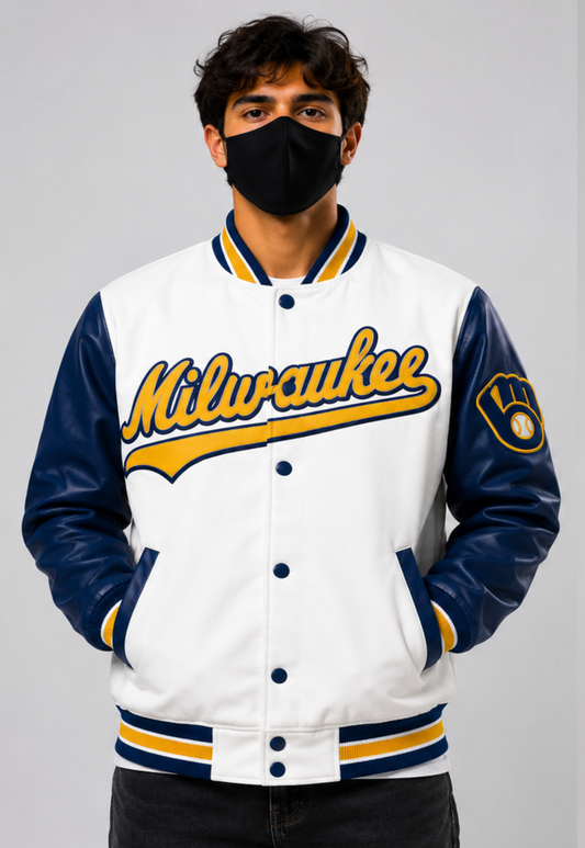 A man wearing a Milwaukee baseball varsity jacket with navy blue leather sleeves, a white body, and yellow and navy striped ribbing, paired with a black face mask against a light gray background.