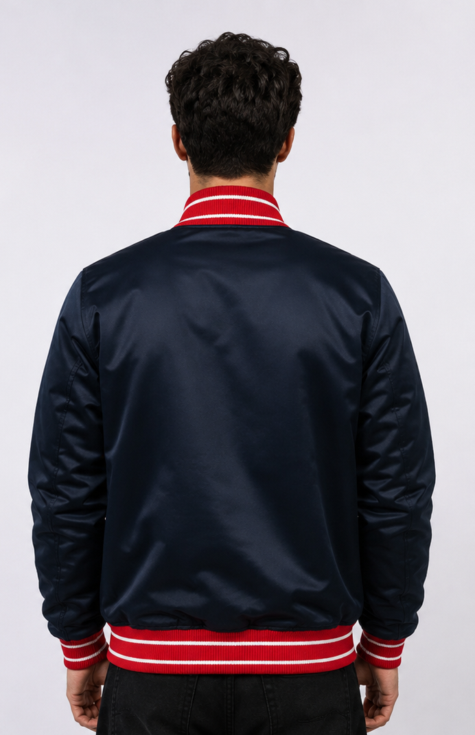 A man viewed from behind wearing a navy blue satin bomber jacket with red and white striped ribbed collar, cuffs, and hem, against a light gray studio background.