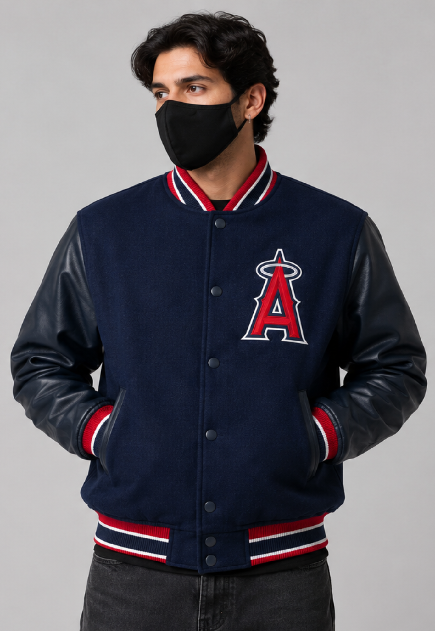 A man wearing a navy blue varsity jacket with faux leather sleeves, featuring the Los Angeles Angels 'A' logo on the chest, red and white striped ribbing, and a black face mask, set against a solid gray background.