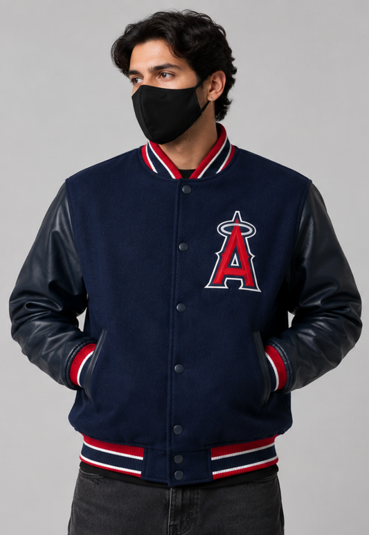 A man wearing a navy blue varsity jacket with faux leather sleeves, featuring the Los Angeles Angels 'A' logo on the chest, red and white striped ribbing, and a black face mask, set against a solid gray background.
