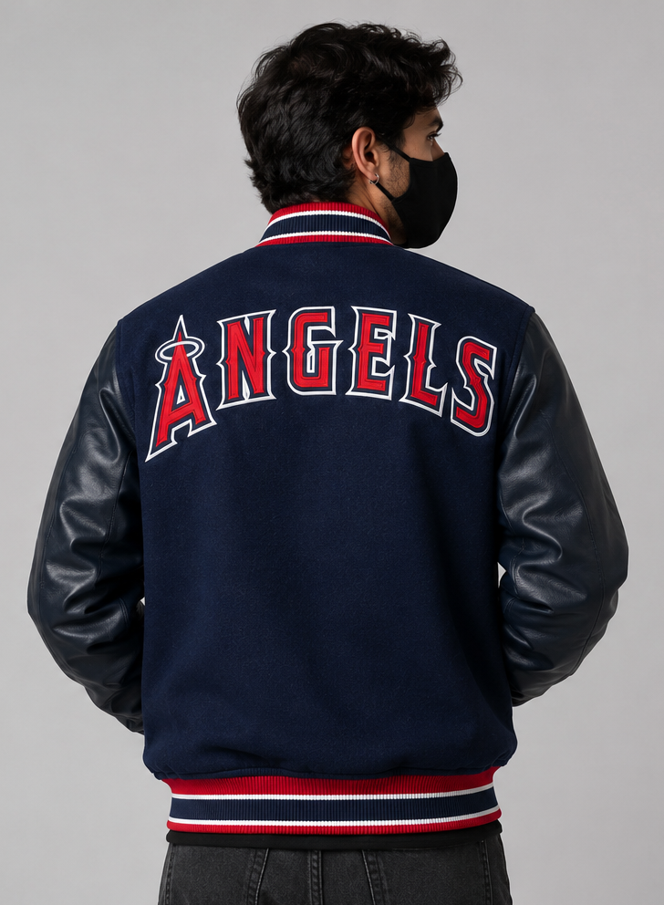 A man viewed from the back wearing a navy blue varsity jacket with faux leather sleeves and 'ANGELS' in red and white across the back, paired with a black face mask against a solid gray background.