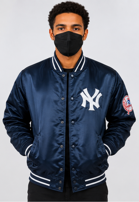 A man wearing a navy blue satin New York Yankees varsity jacket with white ribbed trim and a black face mask, photographed against a light gray studio background.