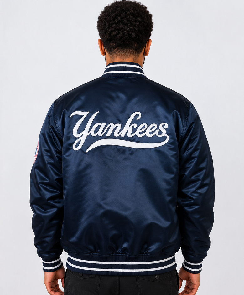 A man viewed from behind wearing a navy blue satin bomber jacket with 'Yankees' written in white script across the back, on a light gray studio background.