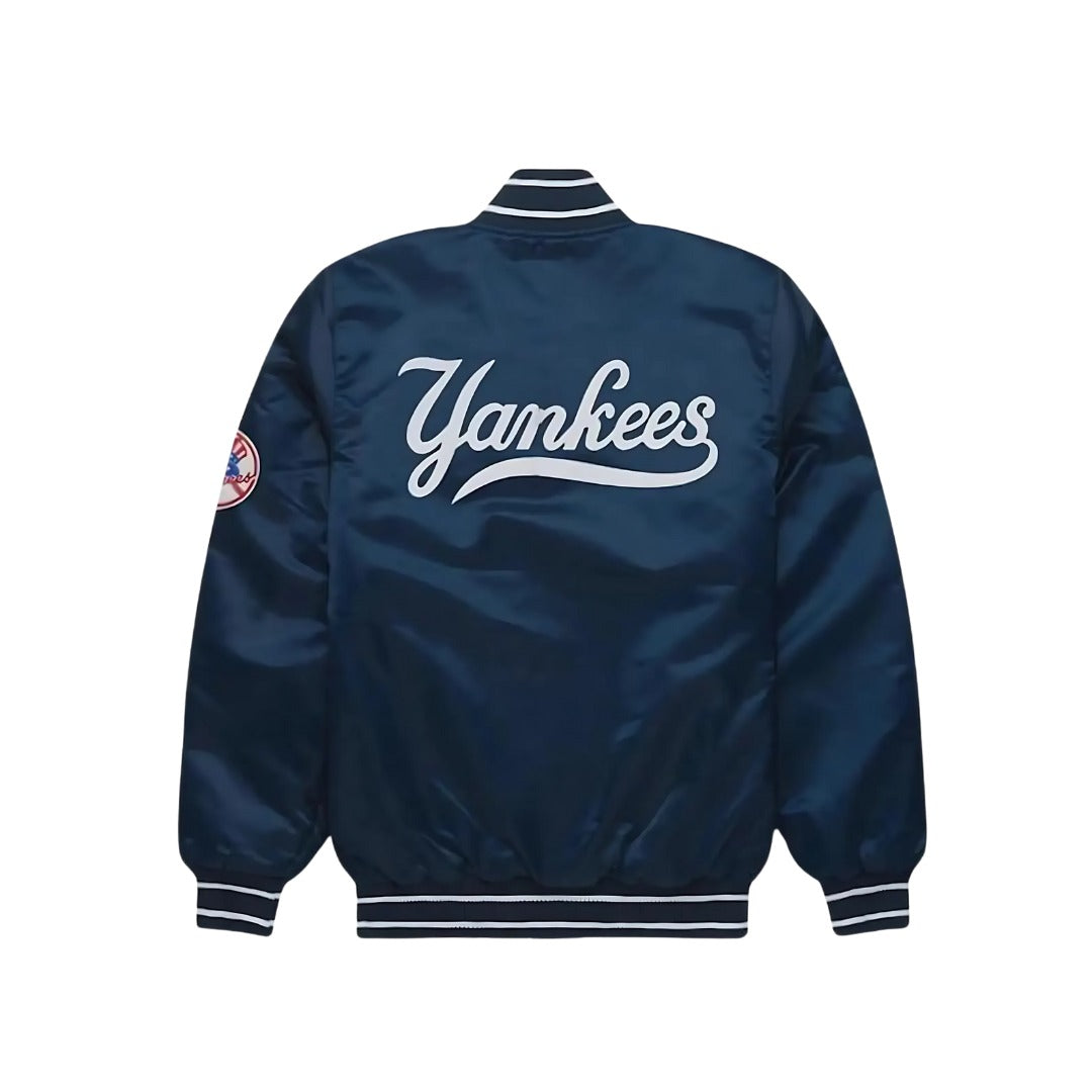 A navy blue satin baseball jacket shown from the back, featuring a large white cursive 'Yankees' logo across the center and a small circular patch on the left sleeve, set against a plain white background.