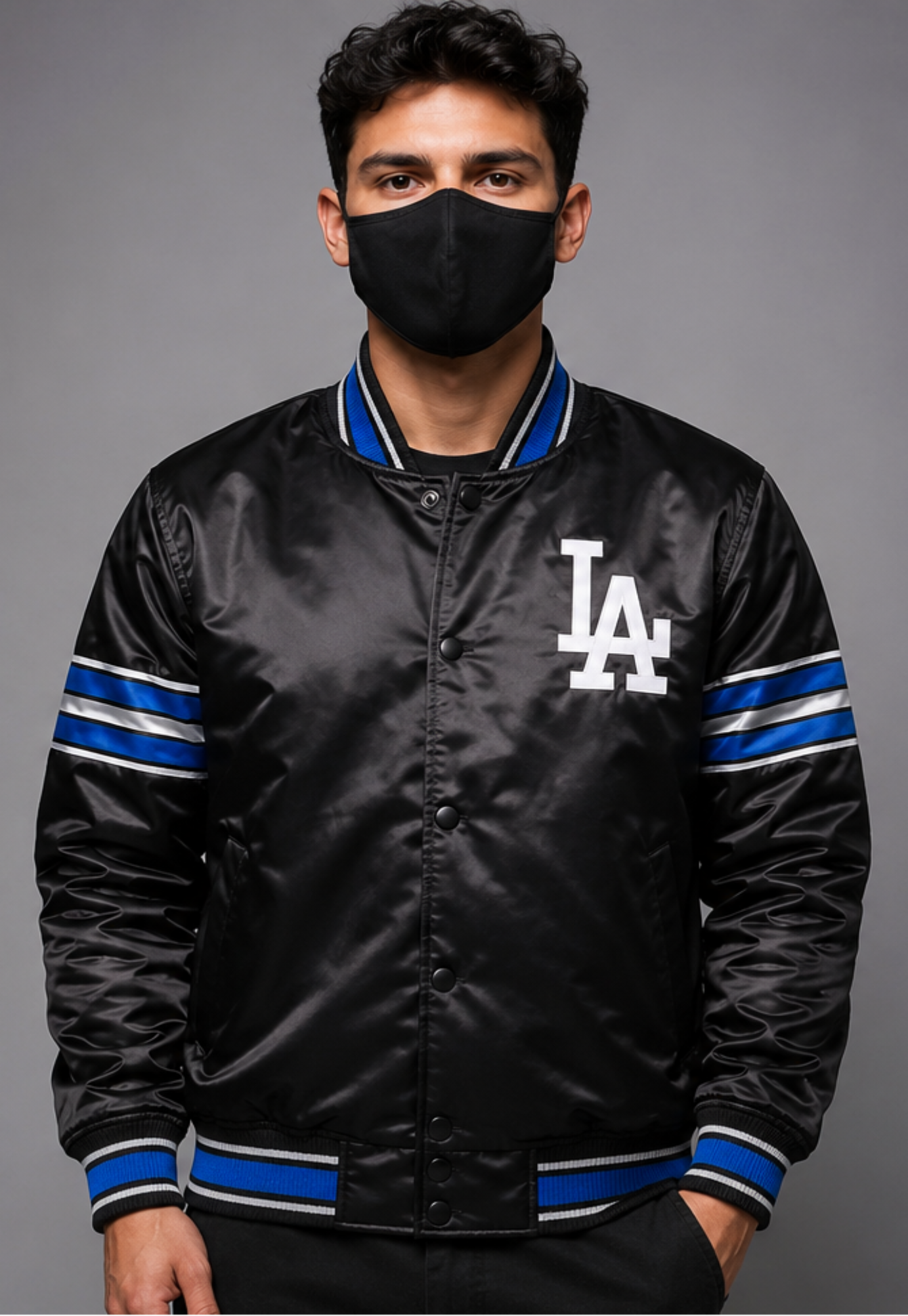 A man wearing a black satin varsity jacket with blue and white striped ribbed trim and a white 'LA' logo on the chest, paired with a black face mask, against a solid gray studio background.