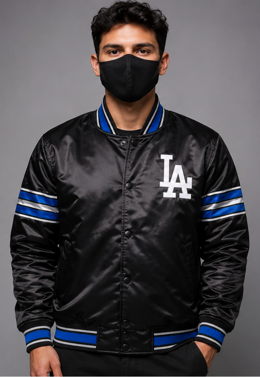 A man wearing a black satin varsity jacket with blue and white striped ribbed trim and a white 'LA' logo on the chest, paired with a black face mask, against a solid gray studio background.