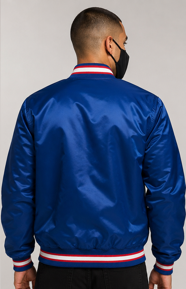 A man viewed from the back wearing a royal blue satin bomber jacket with red and white striped ribbed collar, cuffs, and hem, paired with a black face mask and black pants, against a neutral gray background.