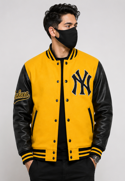 A man wearing a yellow and black New York Yankees varsity jacket with black leather sleeves, a black face mask, and a black shirt, photographed against a light gray studio background.