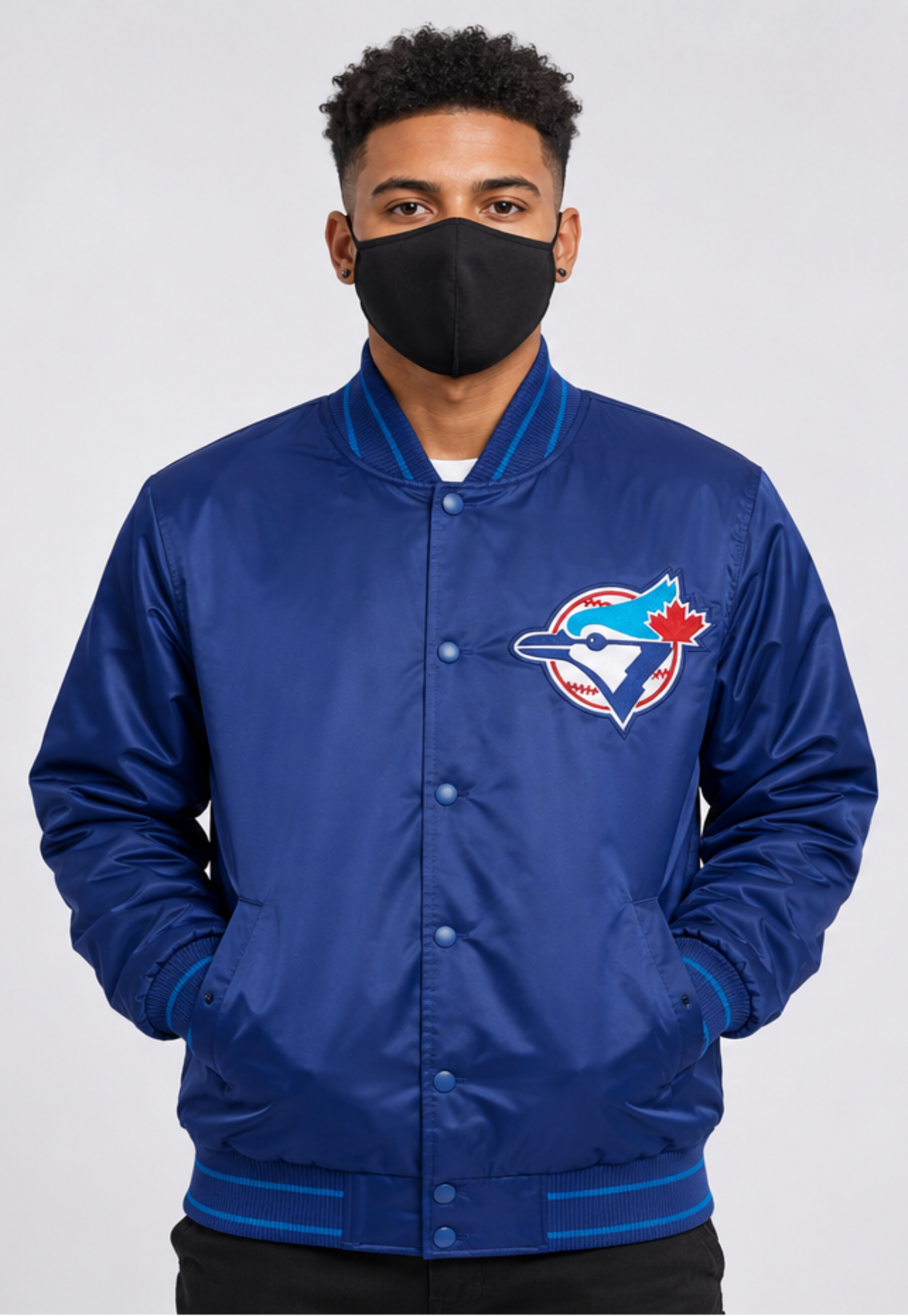 A man wearing a blue Toronto Blue Jays satin bomber jacket with a black face mask, photographed against a light gray studio background.