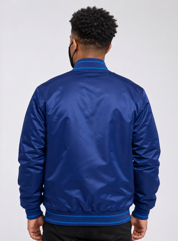 A man viewed from behind wearing a royal blue satin bomber jacket with dark blue ribbed trim on the collar, cuffs, and hem, against a light gray studio background.