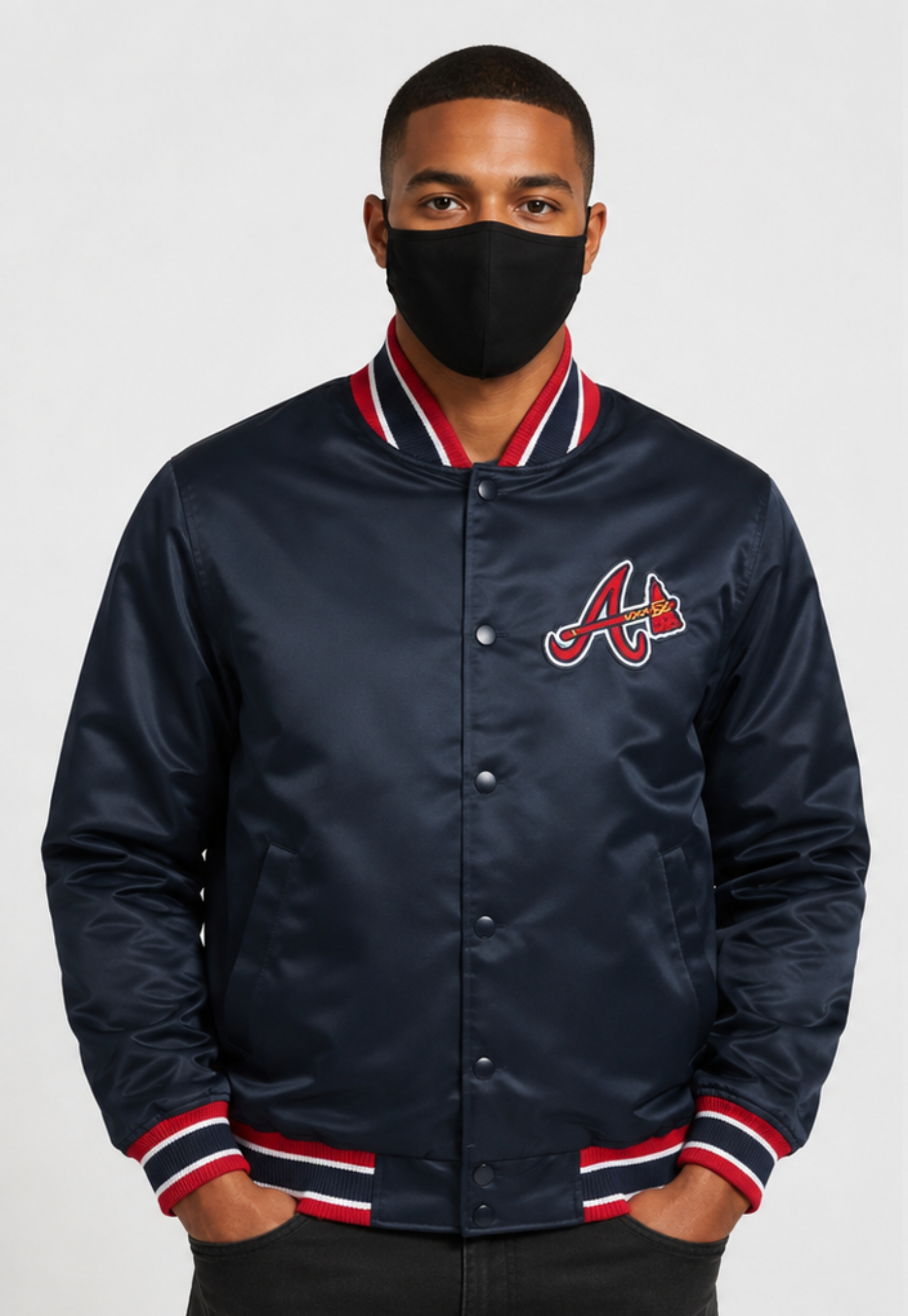 A man wearing a navy blue satin baseball jacket with a red and white striped ribbed collar, cuffs, and hem, featuring the Atlanta Braves 'A' logo on the chest. He is wearing a black face mask and standing against a light gray background.