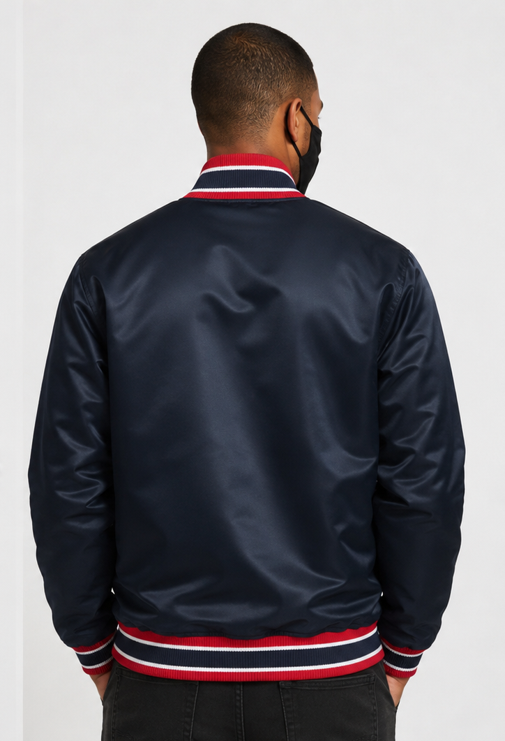 A man viewed from the back wearing a navy satin bomber jacket with red and white striped ribbed collar, cuffs, and hem, paired with a black face mask and dark pants, against a light gray studio background.