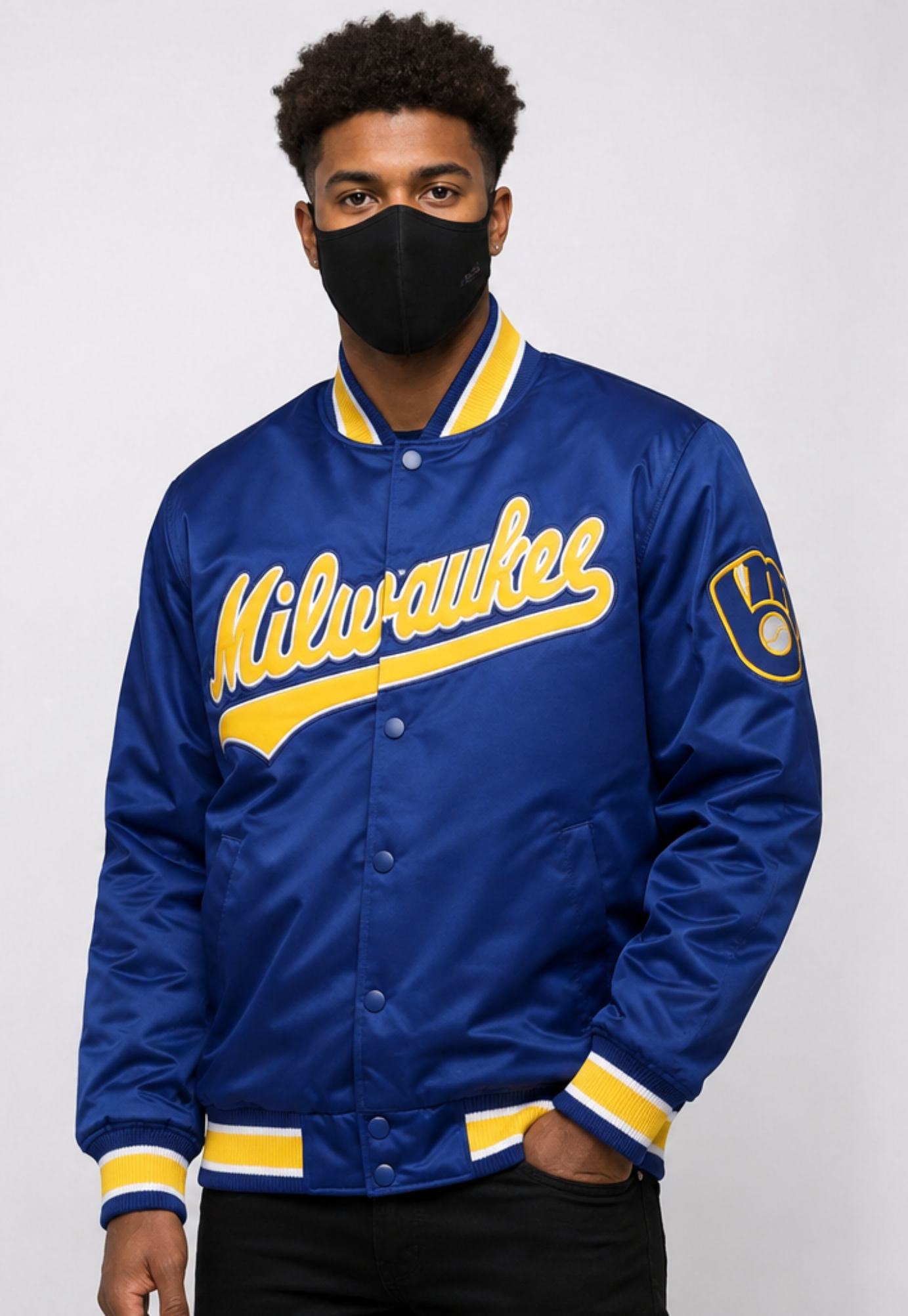 A man wearing a royal blue Milwaukee varsity-style satin jacket with yellow lettering and trim, paired with a black face mask and black pants, against a light gray studio background.