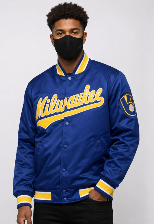 A man wearing a royal blue Milwaukee varsity-style satin jacket with yellow lettering and trim, paired with a black face mask and black pants, against a light gray studio background.