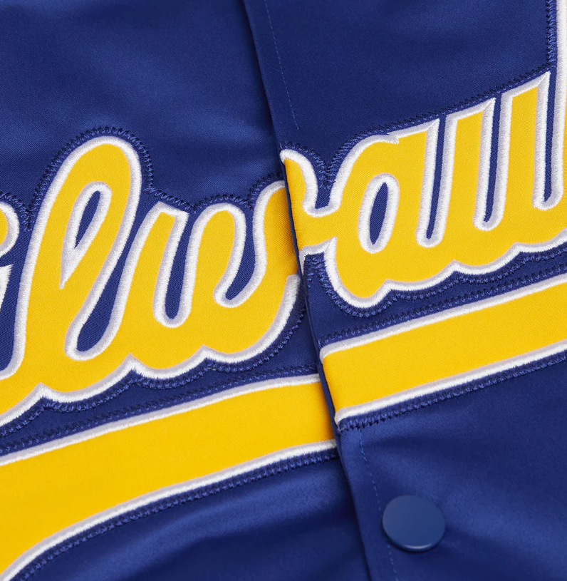 Close-up of a royal blue baseball jacket featuring yellow and white embroidered script lettering across the chest and a matching yellow stripe with white edging.