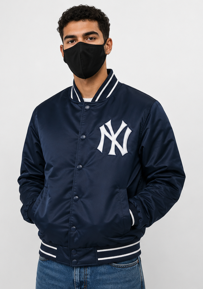 A man wearing a navy blue New York Yankees varsity jacket with a white embroidered NY logo on the chest, paired with a black face mask and blue jeans, against a light gray studio background.